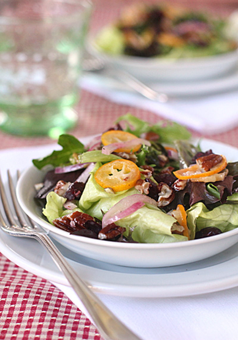 A fresh salad with mixed greens, red onion, dried cranberries, candied pecans, and orange slices is served in a white bowl on a plate, with a fork beside it on a red checkered tablecloth.