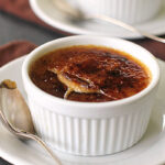 A white ramekin filled with crème brûlée sits on a white plate with a silver spoon. The caramelized sugar top is cracked, revealing the creamy custard beneath. Another ramekin of crème brûlée is visible in the background.