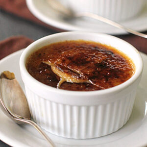 A white ramekin filled with cr&egrave;me br&ucirc;l&eacute;e sits on a white plate with a silver spoon. The caramelized sugar top is cracked, revealing the creamy custard beneath. Another ramekin of cr&egrave;me br&ucirc;l&eacute;e is visible in the background.