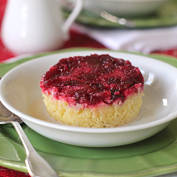 A round dessert with a golden yellow base and a vibrant red fruit topping is served on a white plate with a spoon, placed on a green plate and a red tablecloth. A white pitcher and another plate are in the background.