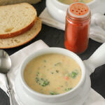 Two white bowls of creamy broccoli soup are on a table, accompanied by slices of bread, a spoon, a white napkin, and a jar of red spice. The soup contains visible broccoli and carrot pieces.