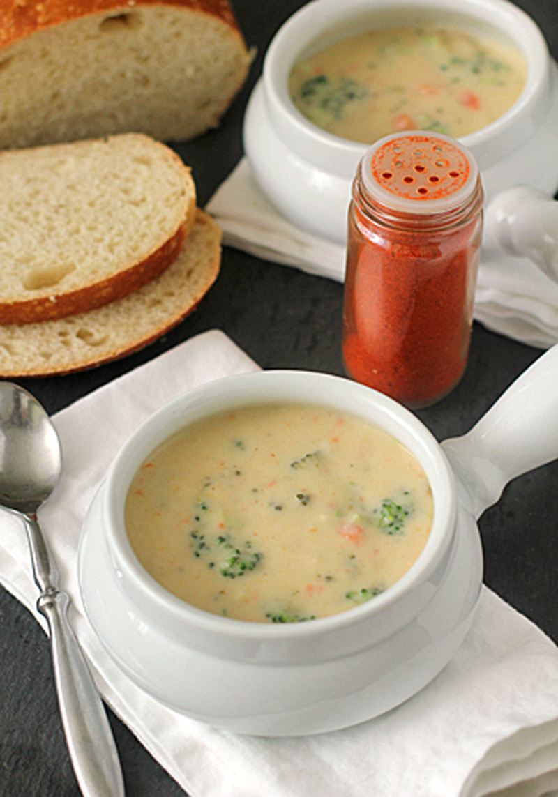 Two white bowls of creamy broccoli soup are on a table, accompanied by slices of bread, a spoon, a white napkin, and a jar of red spice. The soup contains visible broccoli and carrot pieces.