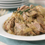 A white plate filled with tender, cooked pork pieces and sliced onions in a light sauce, garnished with a sprig of fresh thyme, set on a blue placemat with stacked plates and cutlery in the background.