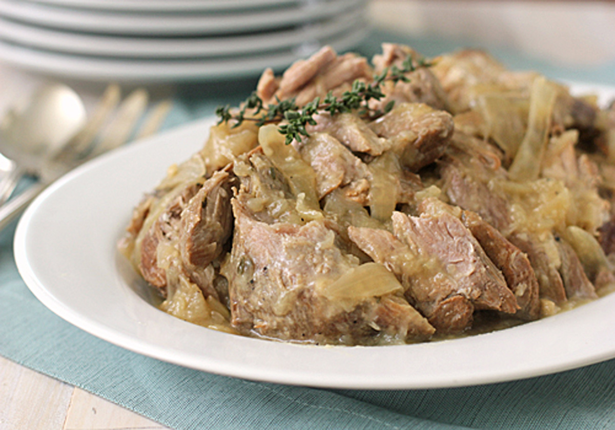 A white plate filled with tender, cooked pork pieces and sliced onions in a light sauce, garnished with a sprig of fresh thyme, set on a blue placemat with stacked plates and cutlery in the background.