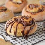 Four chocolate swirl buns rest on a cooling rack, with the bun in front drizzled generously with chocolate sauce. A spoon with extra sauce sits nearby on the wire rack.