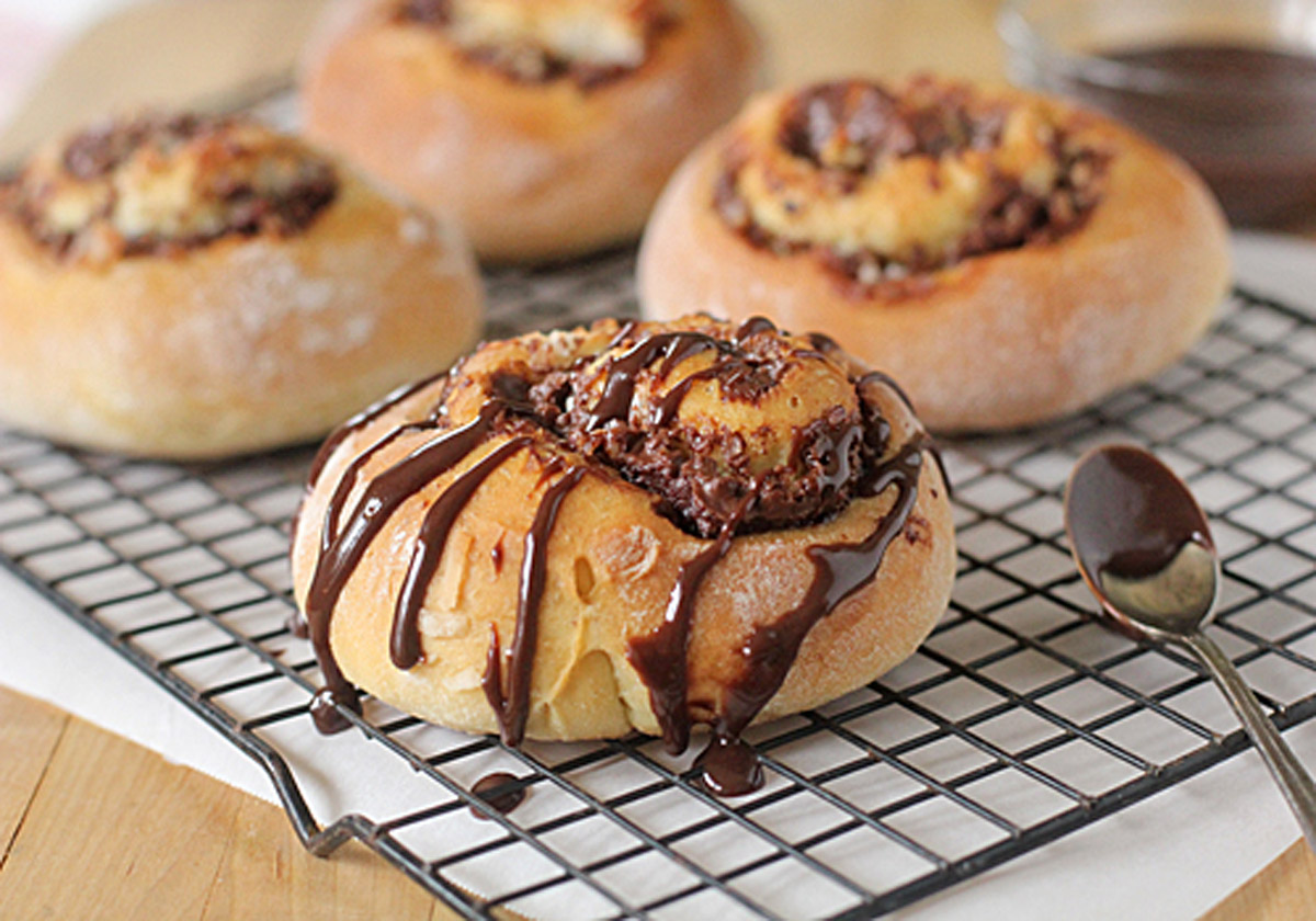 Four chocolate swirl buns rest on a cooling rack, with the bun in front drizzled generously with chocolate sauce. A spoon with extra sauce sits nearby on the wire rack.