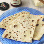 Three pieces of flatbread are arranged on a blue plate. In the background, there is a jar of lingonberry jam with a wooden spoon and a dish of butter with a butter knife.