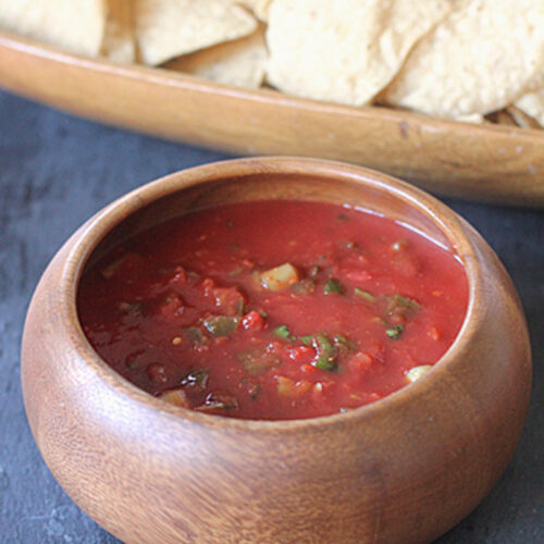 A wooden bowl filled with chunky red salsa sits on a dark surface, with a wooden tray of tortilla chips in the background.