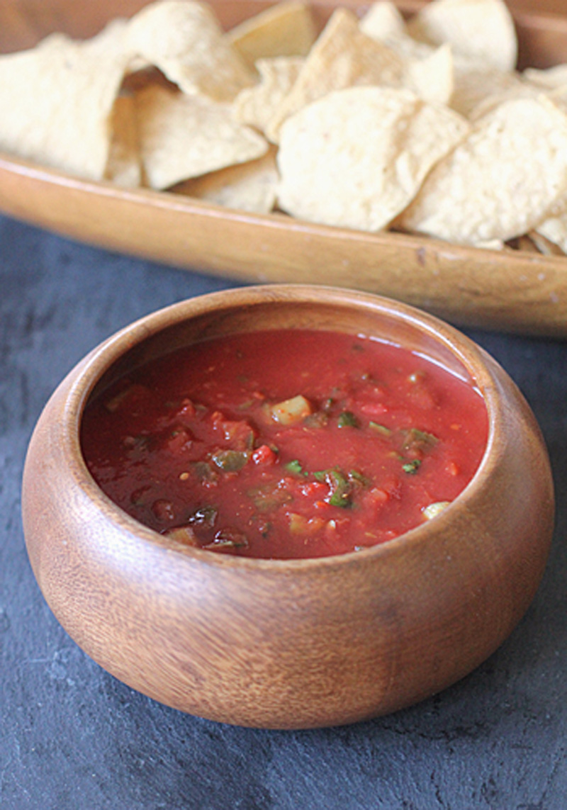 A wooden bowl filled with chunky red salsa sits on a dark surface, with a wooden tray of tortilla chips in the background.
