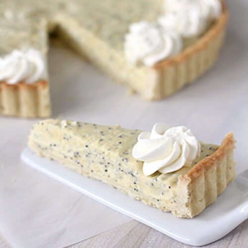 A slice of poppy seed tart topped with whipped cream is served on a small white plate, with the remaining tart and more whipped cream visible in the background.
