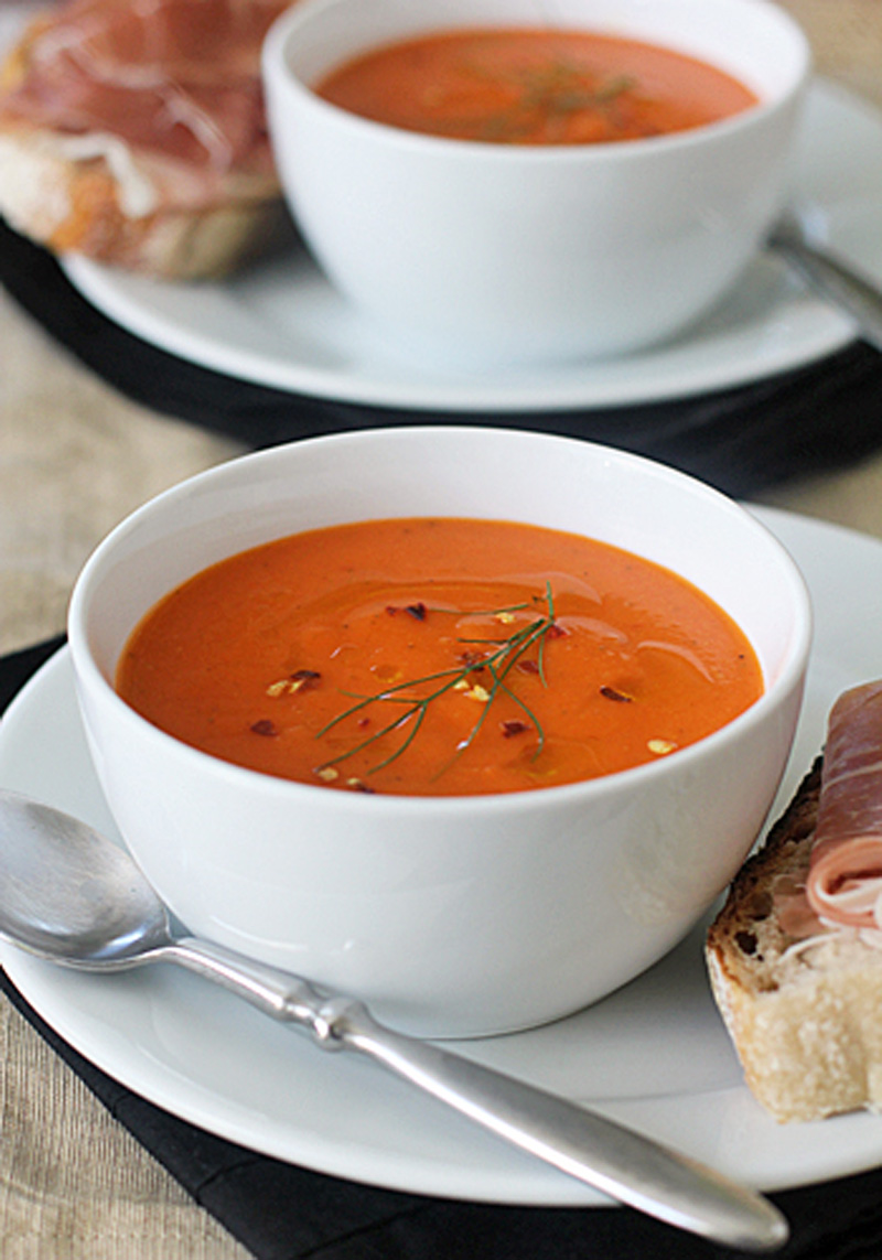 A white bowl of creamy tomato soup garnished with herbs and chili flakes sits on a white plate with a spoon. Next to it is bread topped with prosciutto; another similar setting is blurred in the background.