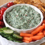 A bowl of creamy spinach dip is surrounded by cherry tomatoes, sliced cucumbers, baby carrots, and crispy crackers, all arranged on a white platter atop a napkin on a wooden table.