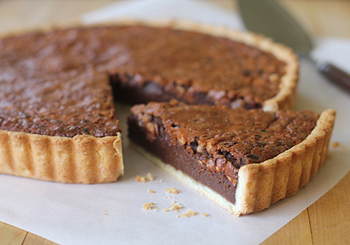 A chocolate tart with a golden crust sits on parchment paper, with one slice cut and slightly pulled out, showing its rich, fudgy filling. A pie server and knife are visible in the background.