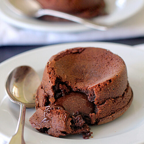 A chocolate lava cake with a gooey center sits on a white plate with a spoon beside it. Another cake and spoon are blurred in the background.