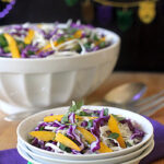 A small bowl of colorful salad with purple cabbage, yellow bell peppers, and greens sits on stacked plates, with a larger bowl of salad and festive Mardi Gras beads in the background.