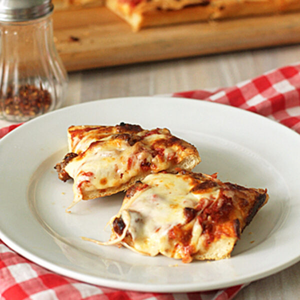 Two slices of cheesy pizza on a white plate, placed on a red and white checkered napkin. A pizza tray and a pepper shaker are in the background.