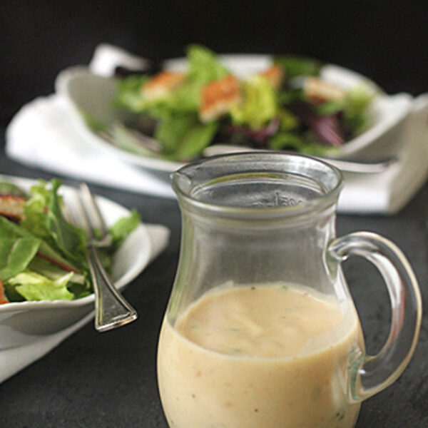 A glass pitcher filled with creamy salad dressing sits on a dark surface, with a fork and plates of fresh green salad in the background.