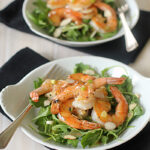 Two white plates with sautéed shrimp on a bed of fresh greens, garnished with sliced almonds. Each plate has a fork and is set on a black napkin atop a light wooden table.