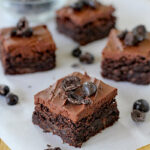 Four pieces of rich, frosted chocolate brownies topped with chocolate-covered cherries are arranged on a white parchment paper, with extra chocolate-covered cherries scattered nearby.