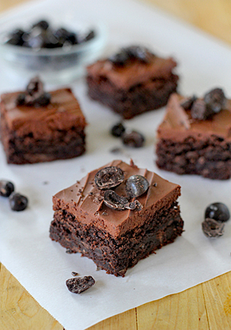 Four pieces of rich, frosted chocolate brownies topped with chocolate-covered cherries are arranged on a white parchment paper, with extra chocolate-covered cherries scattered nearby.