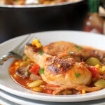 A plate of braised chicken drumsticks served with vegetables like corn, tomatoes, and lima beans in a savory sauce, with a fork on the side and a bottle of hot sauce in the background.