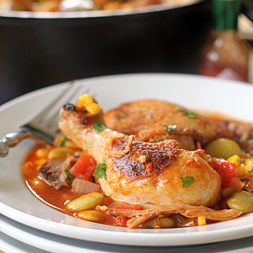 A plate of braised chicken drumsticks served with vegetables like corn, tomatoes, and lima beans in a savory sauce, with a fork on the side and a bottle of hot sauce in the background.