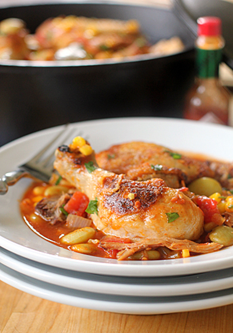 A plate of braised chicken drumsticks served with vegetables like corn, tomatoes, and lima beans in a savory sauce, with a fork on the side and a bottle of hot sauce in the background.