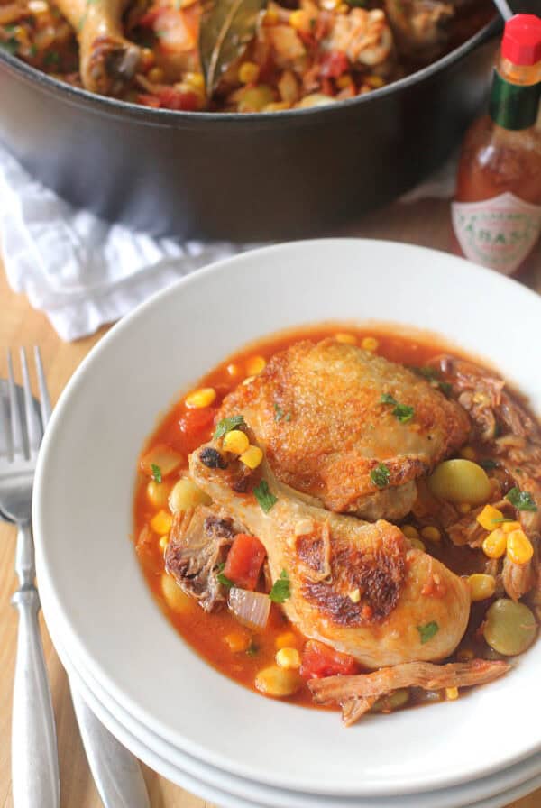 A white bowl of Brunswick Stew, featuring braised chicken with corn, tomatoes, and vegetables in a rich red sauce, sits beside a fork and knife. In the background, a casserole pot filled with the same hearty Southern dish and a bottle of Tabasco sauce can be seen.