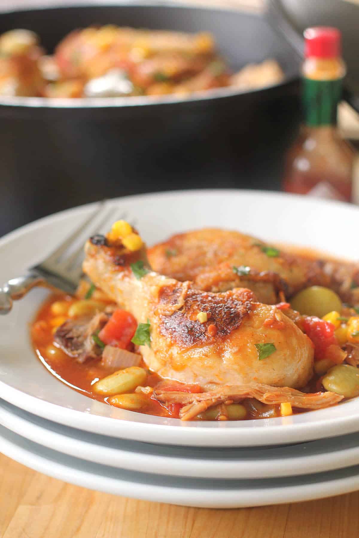 A plate of Brunswick Stew, featuring stewed chicken with corn, lima beans, tomatoes, and herbs, sits on stacked white plates, with a fork and hot sauce bottle nearby.