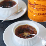 Two white ramekins of crème brûlée, each on a white plate with a spoon. One crème brûlée has a spoon breaking its caramelized sugar top. A Café Du Monde coffee can is in the background.