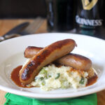 Two browned sausages on creamy mashed potatoes with green herbs, topped with gravy, served on a white plate. In the background, there’s a pint of dark beer and a Guinness bottle. A green napkin is under the plate.
