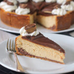 A slice of chocolate-topped cheesecake with a peanut butter cup and whipped cream on top, served on a white plate with a fork, with the rest of the cheesecake in the background.
