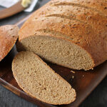A loaf of brown bread on a wooden board, with one slice cut and laid flat; a pat of butter and a knife are blurred in the background.