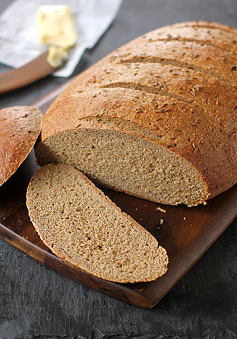A loaf of brown bread on a wooden board, with one slice cut and laid flat; a pat of butter and a knife are blurred in the background.