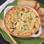 A baked cheesy dip garnished with chopped herbs is served in a round dish on a green plate, with toasted bread slices and a wooden spoon on the side.