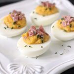 Four deviled eggs topped with chopped ham and sprinkled with herbs are arranged on a white decorative plate.
