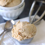 A scoop of light brown ice cream in a metal scoop rests on a white cloth next to two silver spoons, with a bowl containing more ice cream and an ice cream scooper in the background.