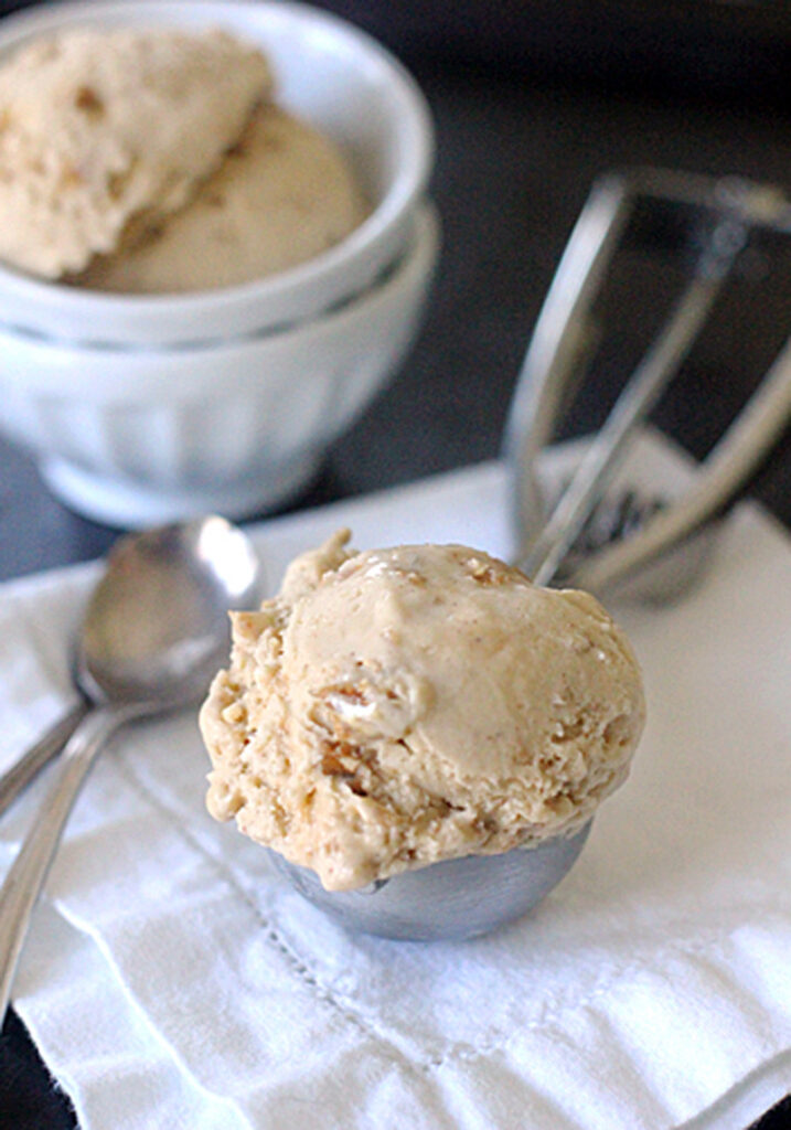 A scoop of light brown ice cream in a metal scoop rests on a white cloth next to two silver spoons, with a bowl containing more ice cream and an ice cream scooper in the background.
