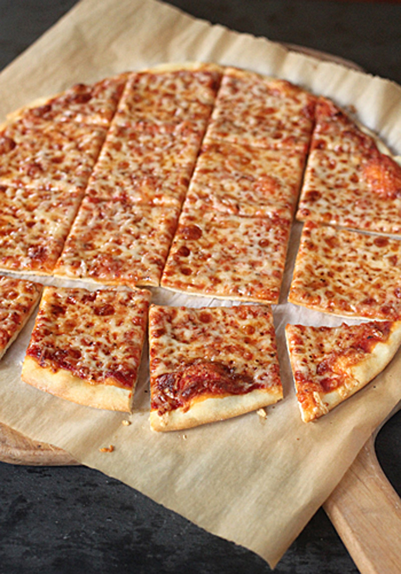 A cheese pizza on parchment paper, cut into rectangular strips and squares, resting on a wooden surface.