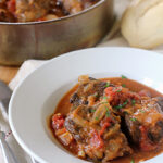 A white bowl filled with braised oxtails in a rich tomato-based sauce, garnished with herbs, sits on a napkin beside a fork and knife. In the background, a pot of oxtails and a loaf of bread are visible.