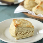 A golden, flaky biscuit sits on a white plate, placed on a light blue cloth. In the background, a basket of more biscuits and a dish of butter are visible.