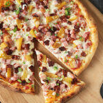 A freshly baked pizza with cheese, ham, bacon, pineapple, and green onions sits on a wooden pizza peel. One slice has been pulled slightly away from the rest of the pizza.