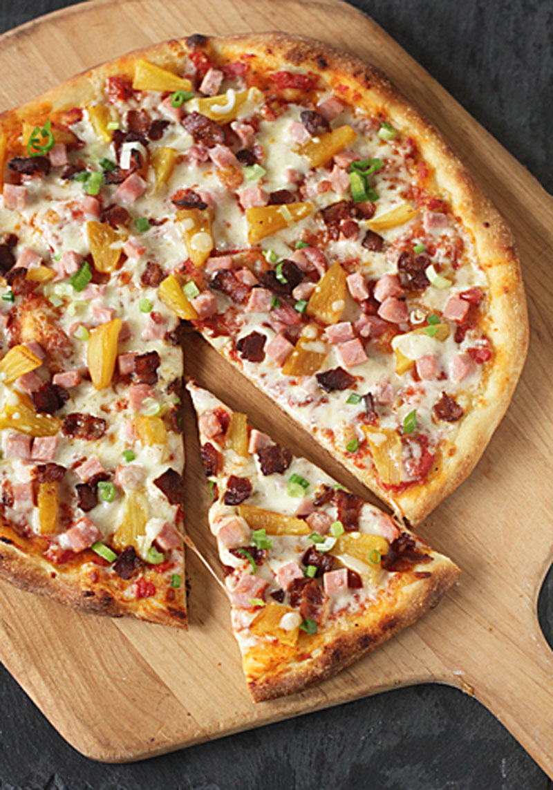 A freshly baked pizza with cheese, ham, bacon, pineapple, and green onions sits on a wooden pizza peel. One slice has been pulled slightly away from the rest of the pizza.