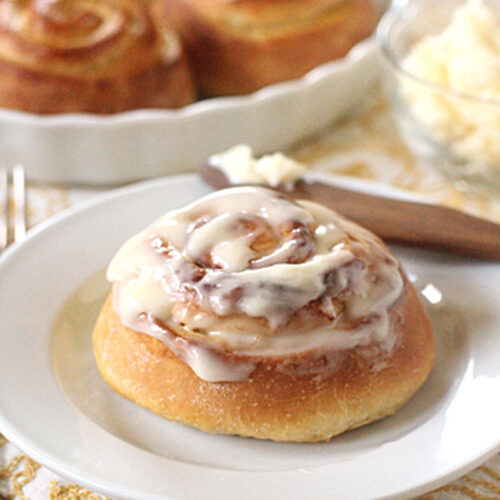 A cinnamon roll topped with creamy icing sits on a white plate. More cinnamon rolls are visible in a dish in the background, along with a bowl of butter and a knife on a patterned cloth.