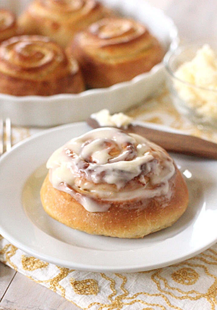 A cinnamon roll topped with creamy icing sits on a white plate. More cinnamon rolls are visible in a dish in the background, along with a bowl of butter and a knife on a patterned cloth.