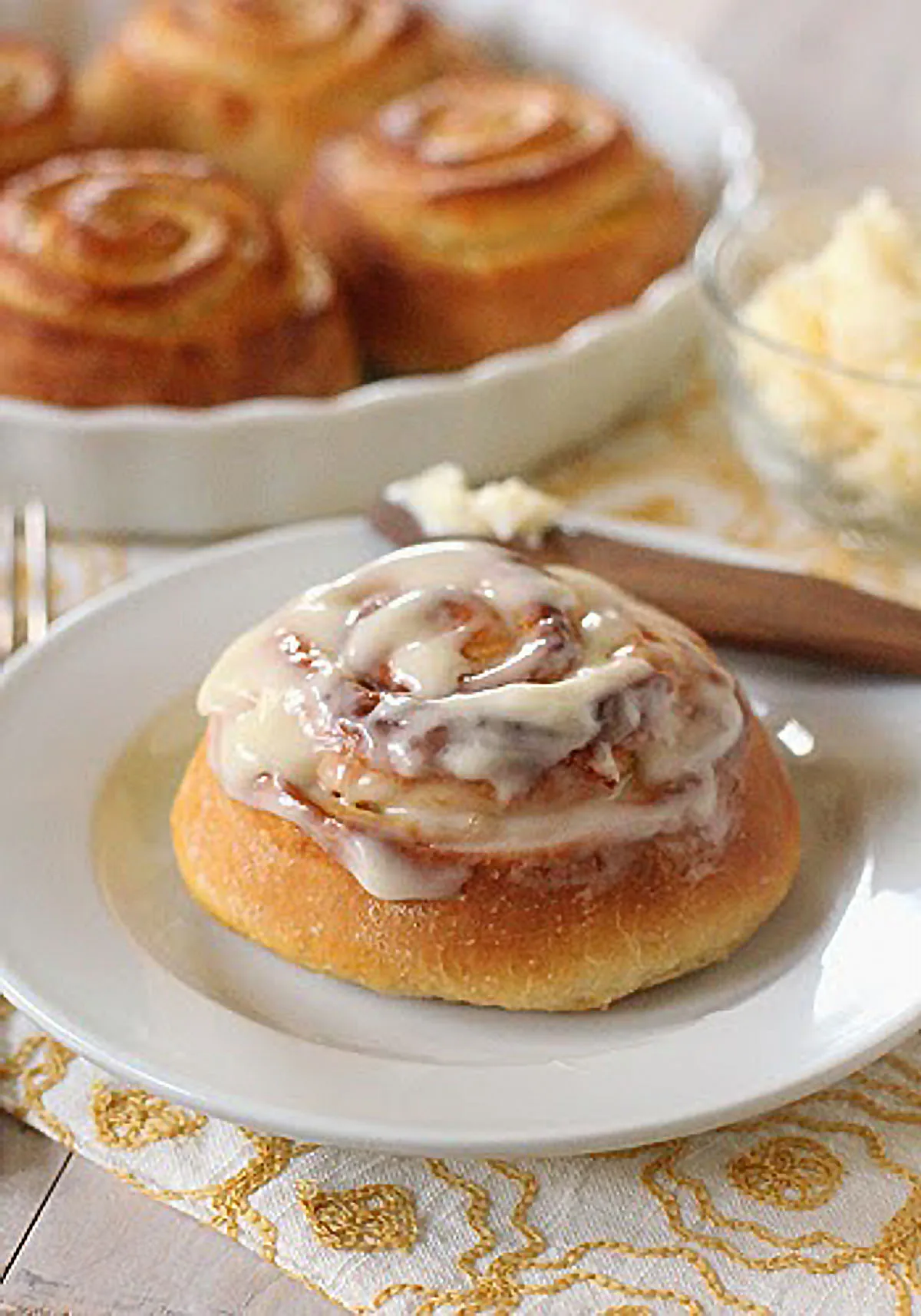 A frosted cinnamon roll sits on a white plate, with more cinnamon rolls on a dish and a bowl of butter in the background on a patterned yellow and white cloth.