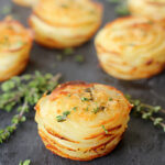 Several stacks of golden, thinly sliced, baked potatoes garnished with fresh herbs are arranged on a dark slate surface, with small sprigs of thyme scattered around.