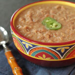 A colorful bowl filled with refried beans topped with sliced jalapeños, sitting on a blue napkin next to a spoon with a wooden handle.