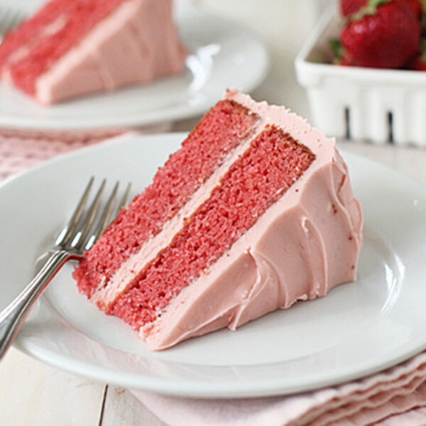 A slice of pink strawberry cake with pink frosting on a white plate, accompanied by a fork. In the background, there is a container of fresh strawberries and another plate with cake.