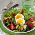 A fresh salad with mixed greens, avocado slices, strawberries, crispy bacon pieces, and a soft-boiled egg, served on a white plate with a green rim. A fork, knife, and clear green glass are nearby.